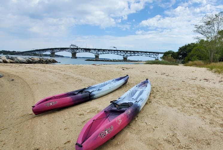 Two pink, purple, light blue kayaks on a sandy beach with a large bridge in the background
