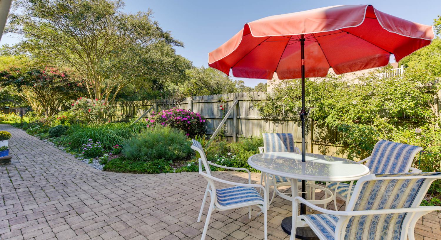 Stone brick patio with white metal patio table and chairs and red umbrella next to large flower garden