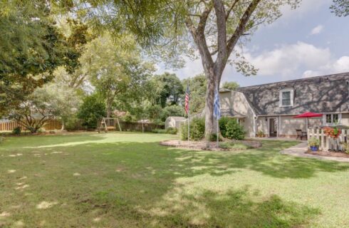 Exterior view of the property painted light taupe with white trim, large backyard with green grass, gardens, and large trees