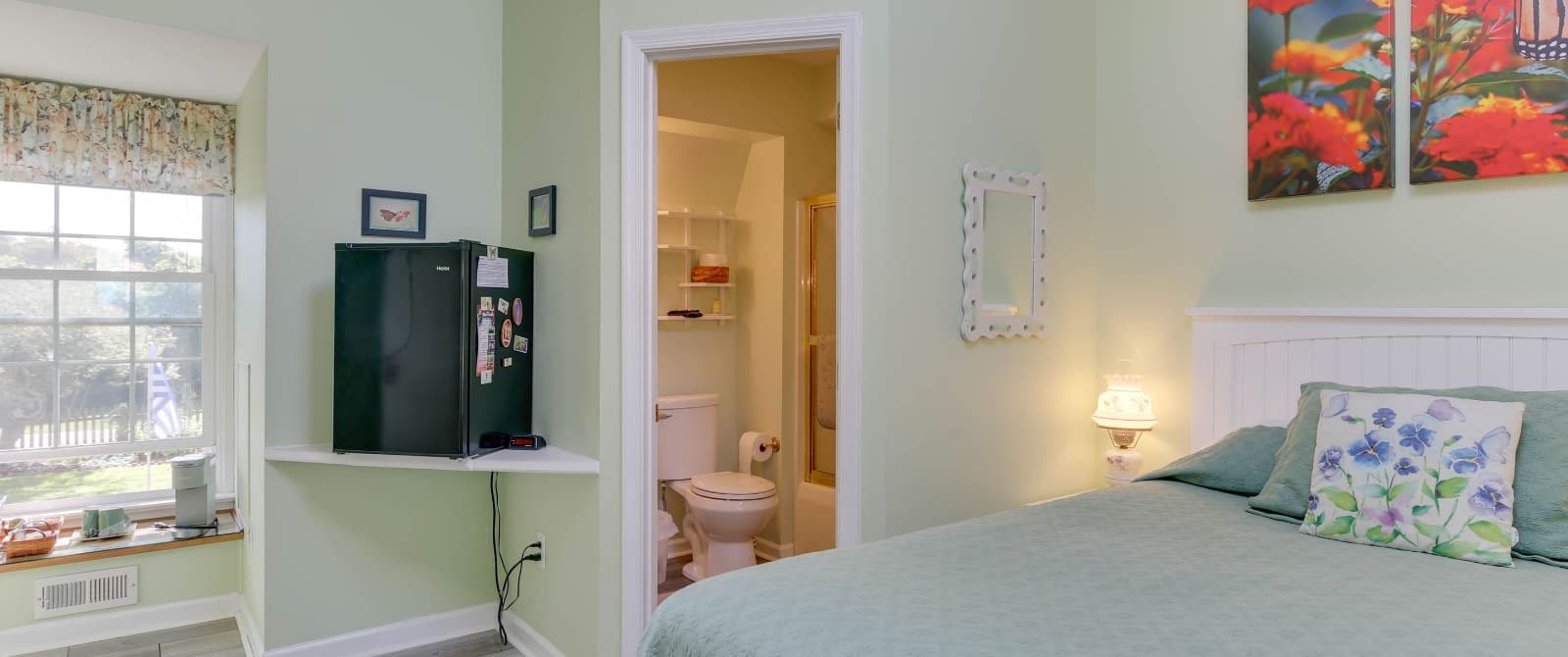 Bedroom with light green walls, hardwood flooring, light green bedding, mini fridge, and view into bathroom