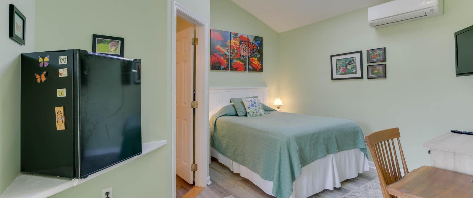 Bedroom with light green walls, hardwood flooring, light green bedding, wood desk and chair, flatscreen TV, and mini fridge