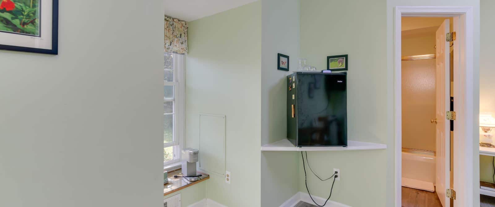 Bedroom with light green walls, hardwood flooring, mini fridge, and view into bathroom
