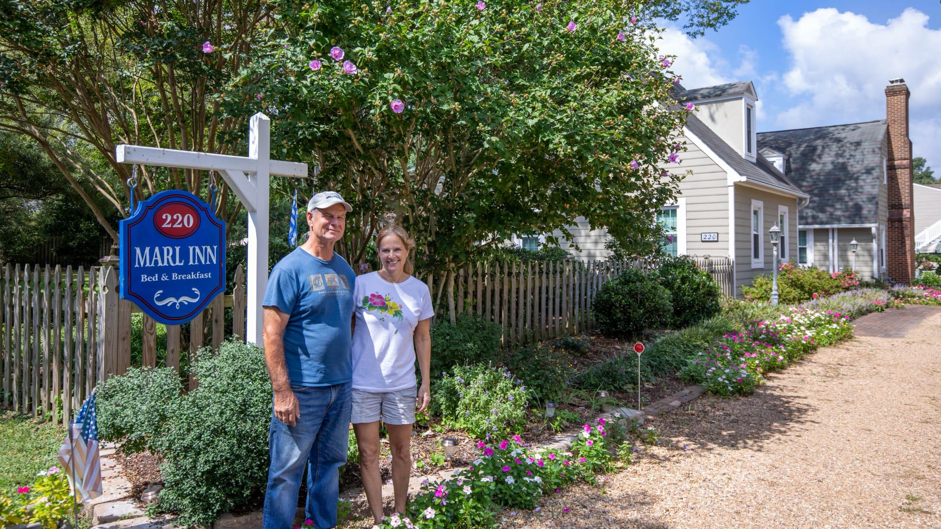 Man and woman standing by Marlk Inn Bed and Breakfast sign with flowers, bushes, and trees in the background