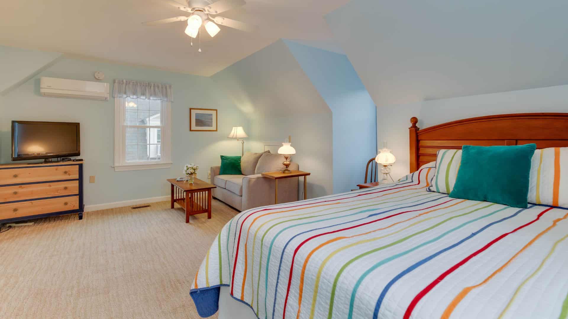 Suite with light blue walls, carpeting, wood headboard, multicolored striped bedding, light gray upholstered sofa, wood coffee table, and flatscreen TV