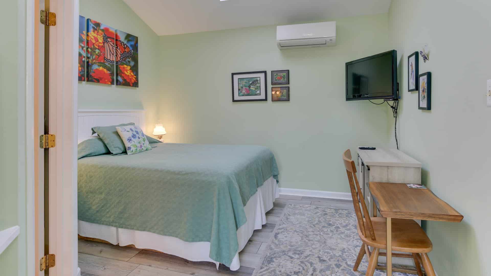 Bedroom with light green walls, hardwood flooring, light green bedding, wood desk and chair, and flatscreen TV