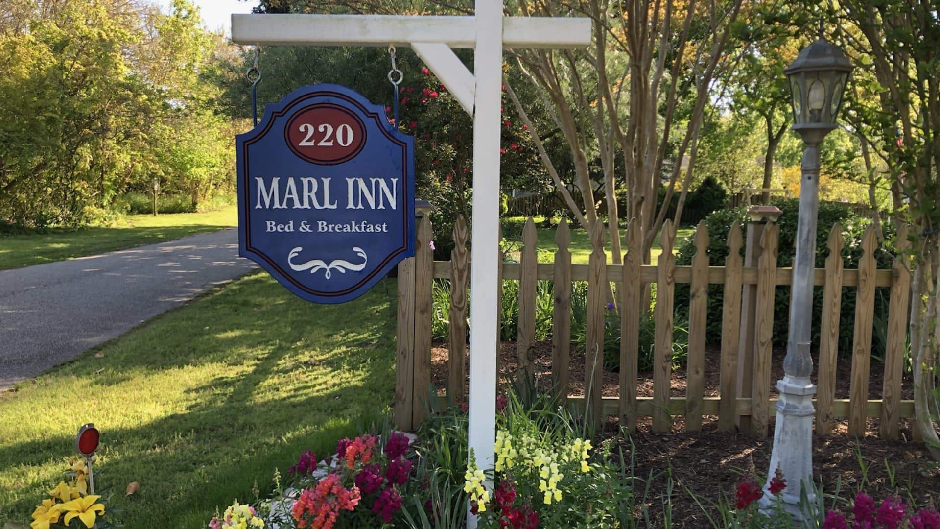 White wooden post holding a blue Marl Inn Bed and Breakfast sign surrounded by flowers and green grass and shrubs