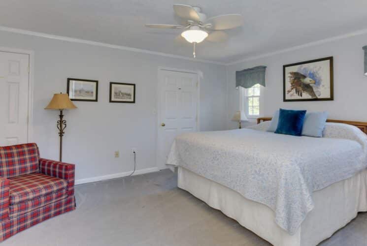 Bedroom with light colored walls, carpeting, light blue and white bedding, red plaid upholstered armchair, and floor lamp