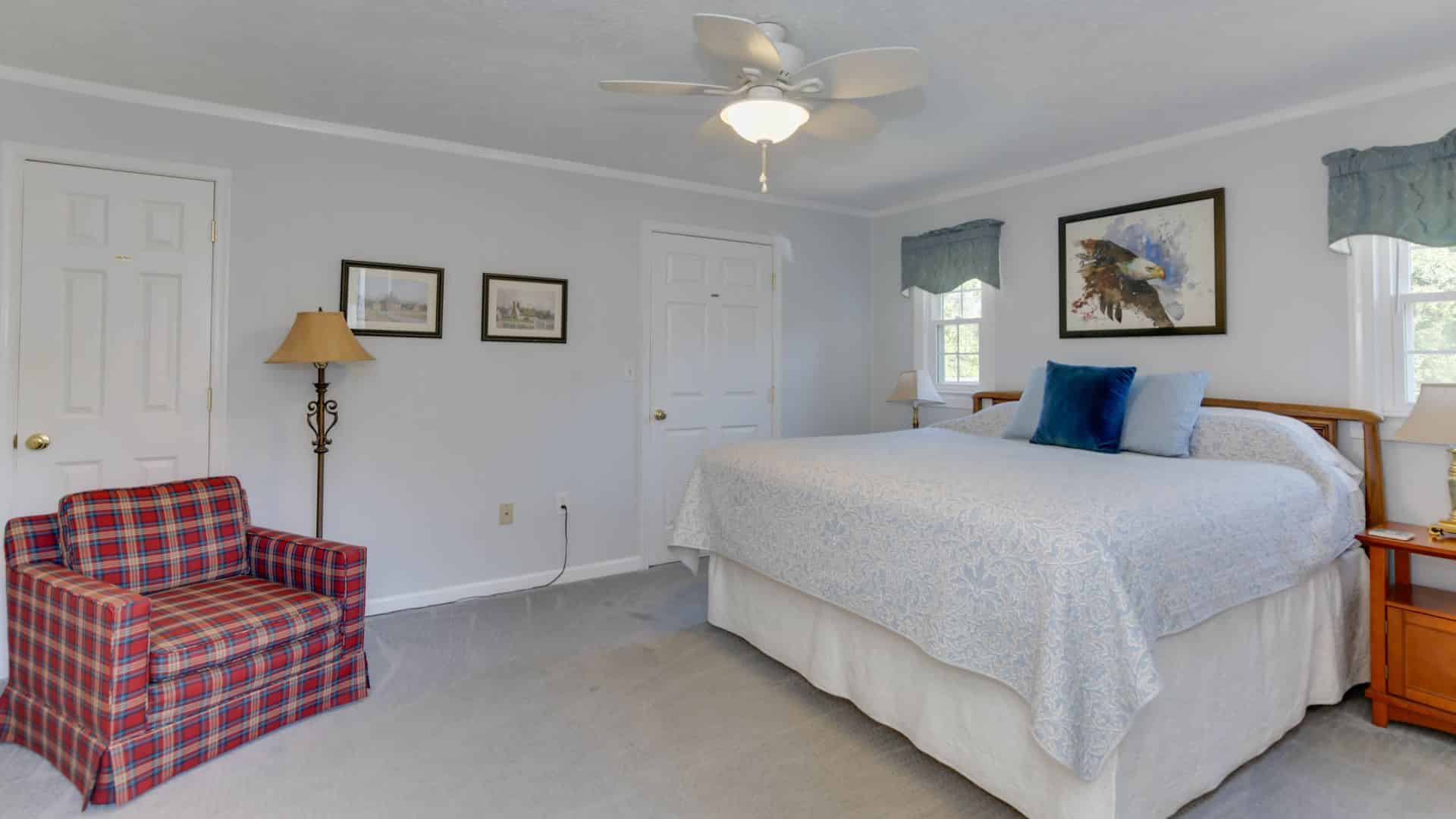 Bedroom with light colored walls, carpeting, light blue and white bedding, red plaid upholstered armchair, and floor lamp