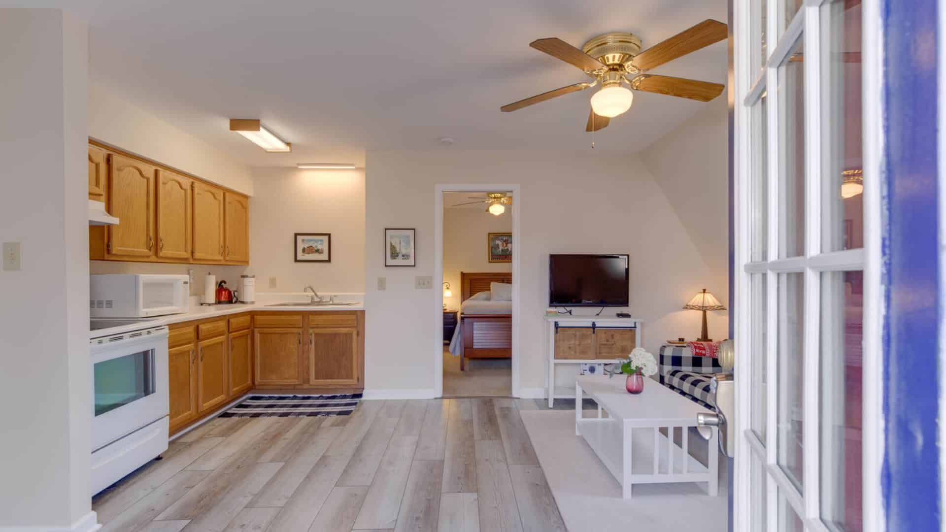 Suite with light colored walls, hardwood flooring, full kitchen, navy and white checked upholstered sofa, white coffee table, flatscreen TV, and view into bedroom